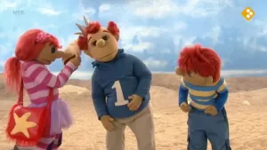 Een foto van de drie poppen, Sassa, Koning Koos en Toto, op het strand. Sassa heeft een schelp in haar hand. 