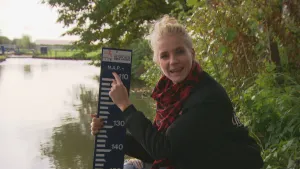 Foto van presentator die staat bij een waterpeilmeter langs een rivier.
