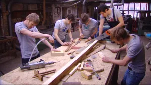 Foto van aantal mensen die aan grote tafel hout bewerken met een zaag etc.