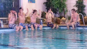 Foto van aantal jongens die op de rand van het zwembad zitten met de benen in het water, bewegen met hun armen.