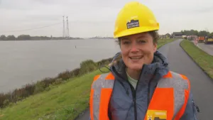 Foto van presentator in werkkleding en met helm op een dijk met weg langs grote waterplas/rivier.