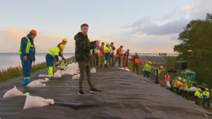 Foto van een crisisoefening overstroming, veel mensen met werkkleding aan die staan op een dijk langs het water.
