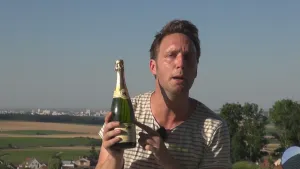Foto van presentator die buiten met weilanden op de achtergrond staat met een fles Champagne in zijn handen.