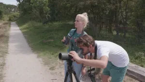 Foto van presentator die staat langs een pad in de natuur bij een fotograaf met fotocamera op statief.
