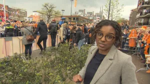Foto van presentator die staat bij een drukke straat in de stad, veel met Oranjekleding aan, tijdens bezoek van de Koning..