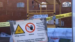 Foto van een gebouw met ervoor afzetlint met waarschuwingsbord voor giftige stoffen, asbest, er aanwezig.