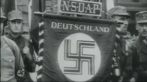 Oude foto, zwart-wit, van een Nazi-vlag met hakenkruis erop, erboven de tekst "Deutschland", weer daarboven "NSDAP", erachter staan aantal nazi's.