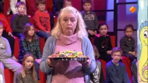 Foto van een oudere vrouw die een schaal met allerlei snoep erop draagt in de studio van Snugger, veel publiek op de achtergrond.