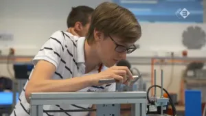 Foto van een jongen, leerling, die in klas onderdeel van een metalen apparaat opmeet met soort liniaal.