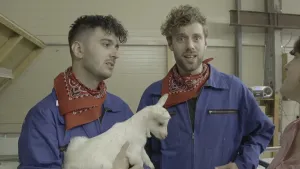 Foto van twee mannen, één van hen houdt een jong bokje, vast.