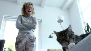 Foto van vrouw die in een huiskamer staat en nog deel van een kat te zien, de kop vooral, foto beetje van onderaf genomen.