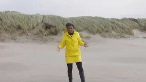 Foto van presentator met gele regenjas aan die staat op het strand, op de achtergrond duinen te zien.