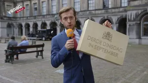 Foto van presentator met microfoon die op plein in de stad koffertje laat zien waarop staat "derde dinsdag in september".
