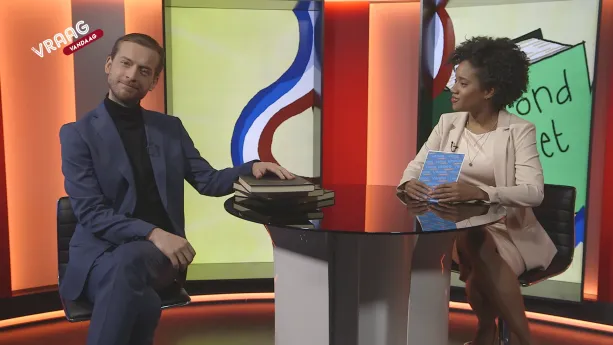 Foto van presentator en man met stapel boeken voor zich die aan een tafeltje zitten in de studio, geschilderde Nederlands vlag op de achtergrond.