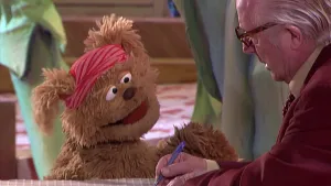 Foto van Sesamstraat met Tommie, pop, in beeld en de presentator die iets schrijft in het vriendenboekje van Tommie.