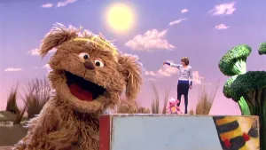 Foto van Sesamstraat waarbij je Tommie, een pop, ziet die zingt, op de achtergrond presentator en mooi blauwe hemel met zon.