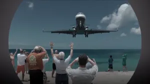 Foto van aantal toeristen op het strand die zwaaien naar een vliegtuig boven de zee dat bijna gaat landen.