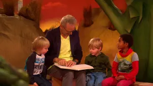 Foto van Sesamstraat waarop een man uit een boek voorleest aan aantal jonge kinderen die bij hem zitten.