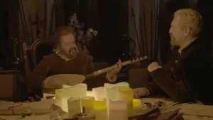 Foto van twee mannen verkleed uit oudere tijd zitten aan tafel met kaarsen erop, één van hen speelt op een luit.