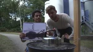 Foto van twee mannen die staan bij een fluitketel die op een rooster staat, één van hen houdt een foto van een grote fabriek vast.