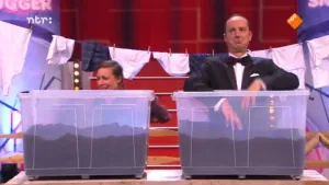 Foto van de presentator van Snugger in de studio, bij hem staat een vrouw, voor hun twee grote bakken met iets van aarde erin, achter hun hangt een waslijn met was.