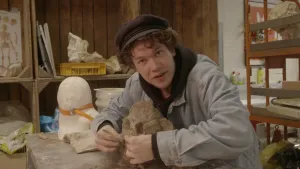 Foto van presentator die zit aan tafel in een atelier, bij hem staat een geboetseerd beeld.