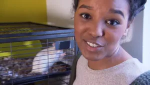 Foto van presentator, je ziet haar gezicht, die staat bij een kooi met een witte rat erin.