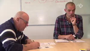 Foto van twee mannen die aan een tafel zitten in een klaslokaal, de ene schrijft, de andere niet.