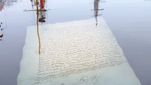 Foto van een oud archiefstuk, vel papier met geschreven tekst, die drijft in het water, in het water spiegelen masten van schepen.