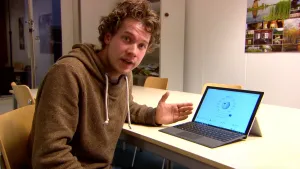 Foto van presentator die zit aan een tafel achter een laptop, hij vertelt iets erover.