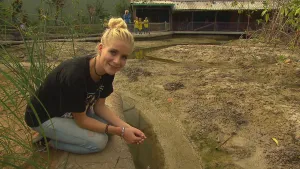 Foto van presentator die gehurkt zit bij een stuk natuurlijke zandgrond met lage begroeiing.
