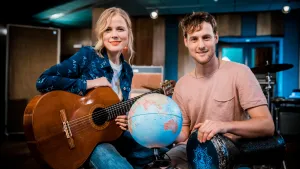 Foto van Ilse de Lange met gitaar in haar handen die zit bij presentator, voor hun staat een wereldbol.
