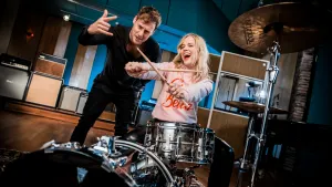 Foto van presentator en Ilse de Lange, zij speelt op de trommel van een drumstel.