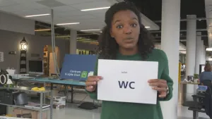 Foto van presentator die in grote bedrijfsruimte staat en een wit vel papier vasthoudt met in grote zwarte letters "WC" erop.