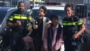 Foto van presentator met microfoon die twee politie-agenten interviewt, op de achtergrond staat een politieauto.