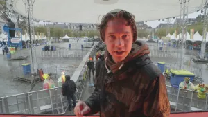 Foto van presentator met op de achtergrond een festivalterrein, momenteel leeg, mensen zijn daar aan het werk.