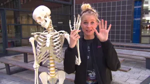 Foto van presentator die staat naast een namaakskelet, geraamte, laat er de hand van zien.