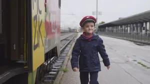 Foto van een jongetje verkleed als conducteur met rode pet op die naast een trein op het perron loopt.