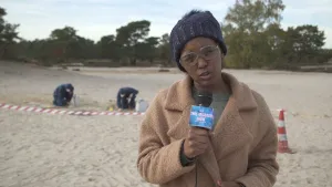 Foto van presentator die staat bij een zandverstuiving, zandduin, aantal personen achter hem doen er onderzoek.