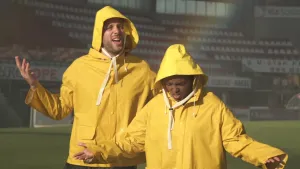 Foto van twee mannen in gele regenpakken met capuchons op die in de regen in een leeg stadion staan.