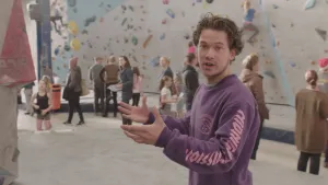 Foto van presentator die staat in een boulderhal met veel mensen en kinderen, sommigen aan het boulderen tegen hoge muur.