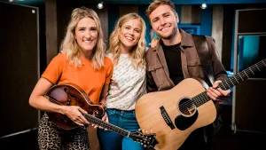 Foto van presentator met Ilse DeLange bij andere vrouw en man, Suzan en Freek, ze houden twee gitaren vast.