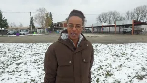 Foto van presentator die staat in besneeuwd grasveld, op de achtergrond gebouwen van een vluchtelingenkamp.