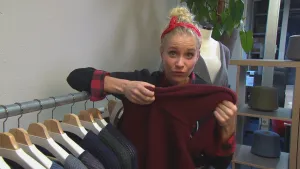 Foto van presentator bij een rek met kleding eraan, ze laat een donkerrode trui zien.