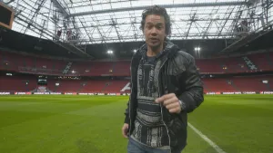 Foto van presentator in een groot voetbalstadion zonder publiek, staat op het gras van het veld.