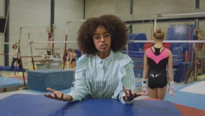 Foto van presentator in een gymnastiekzaal met turntoestellen en meisjes die er trainen.