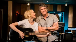 Foto van Ronnie Flex die staat en Ilse de Lange die zit en op een gitaar speelt.