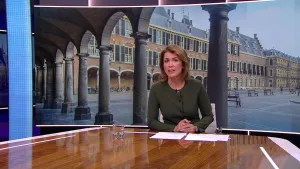 Foto van presentator die zit aan een grote tafel, achter haar foto van het Binnenhof te Den Haag.