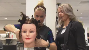 Foto van twee vrouwen die staan bij een namaak vrouwen mensenhoofd, één van hen knipt de haren ervan.