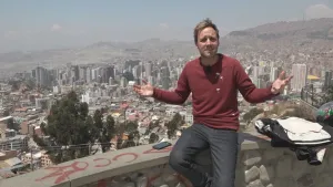 Foto van presentator die zit op een muurtje met erachter heel in de diepte een grote stad, La Paz in Bolivia.